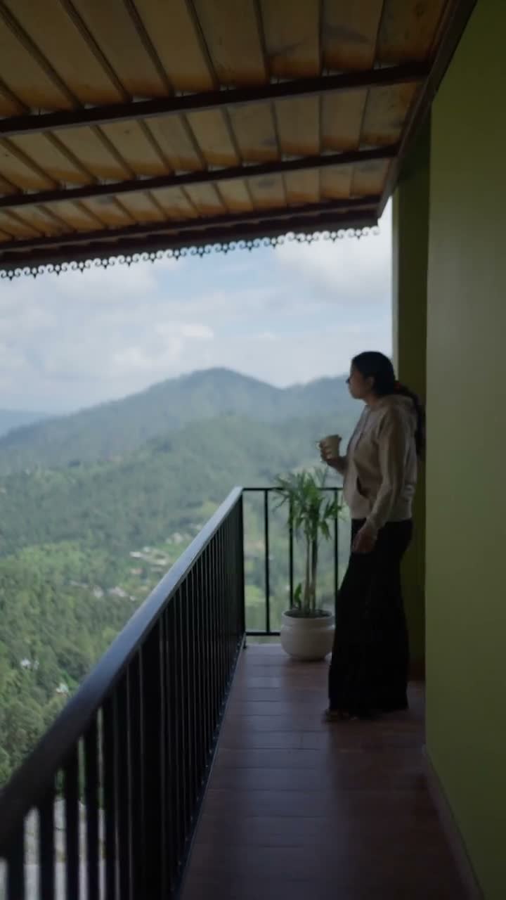 A guest stands at the veranda railing with a mug, looking out over layered green ridges